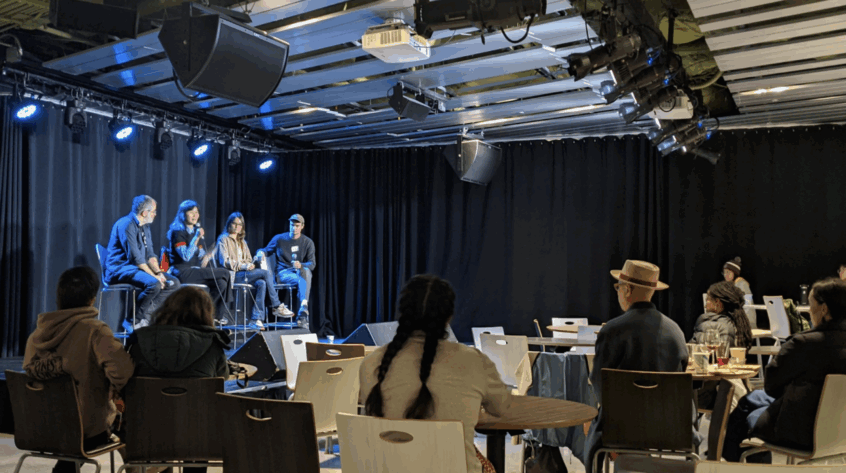 Four individuals sit on a stage, as one speaks into a microphone. They sit in front of a small crowd.